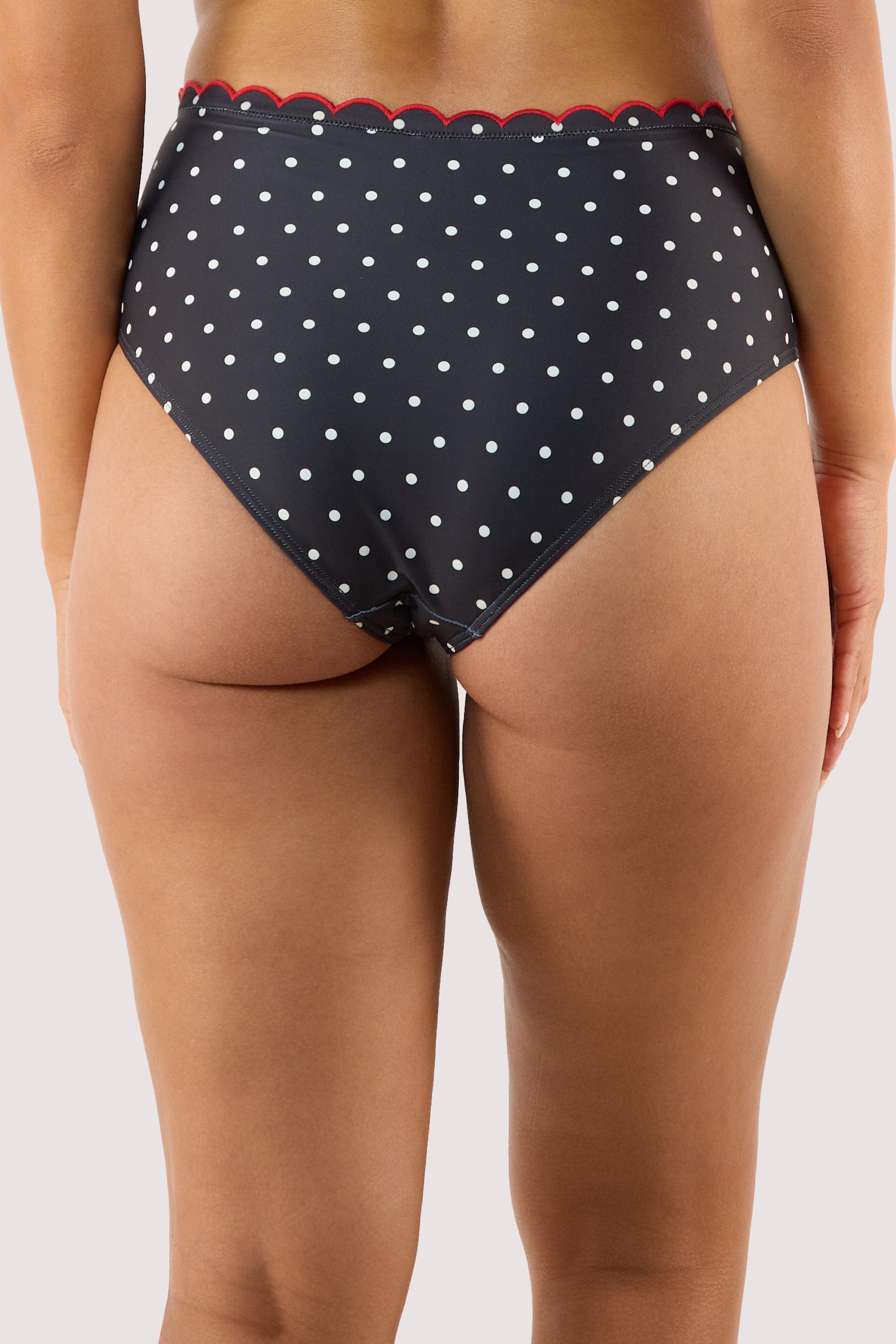 Model Back view in black and white polka dot high-waist bikini briefs with red scalloped edges, showcasing retro style.