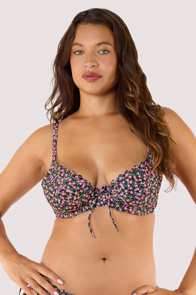 Model in a black ditsy-floral bikini top with scalloped edges and tie detail, posing confidently against a light background.