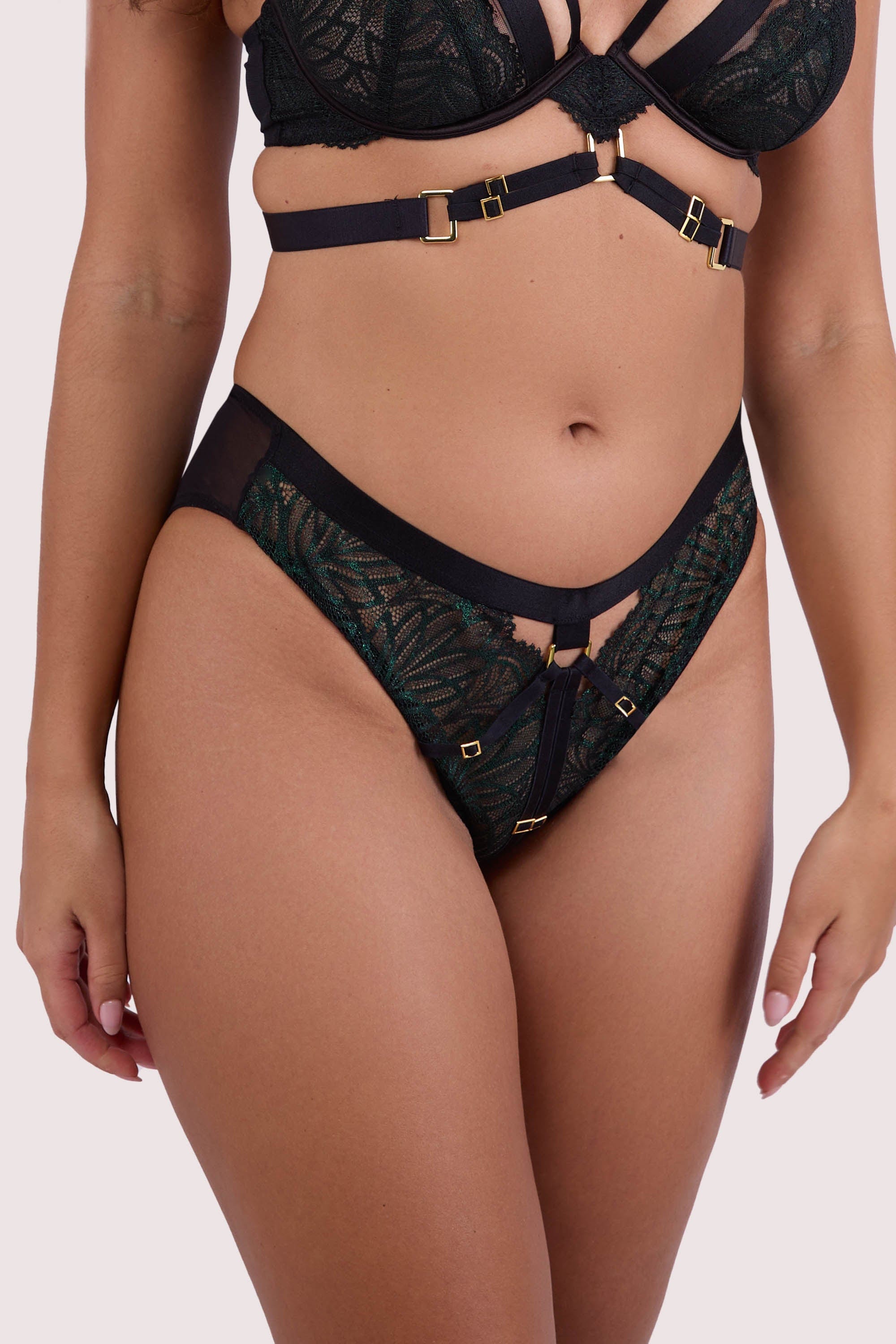 Model wears dark green wet look lace thong with black harness straps and gold-tone hardware