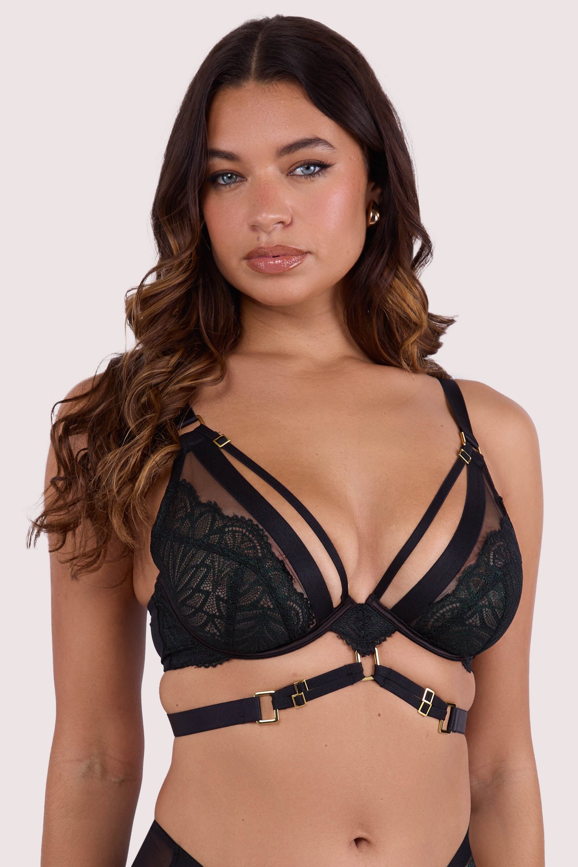 Model wears dark green wet-look plunge bra with underwire cups, strappy harness design, and gold buckle detailing