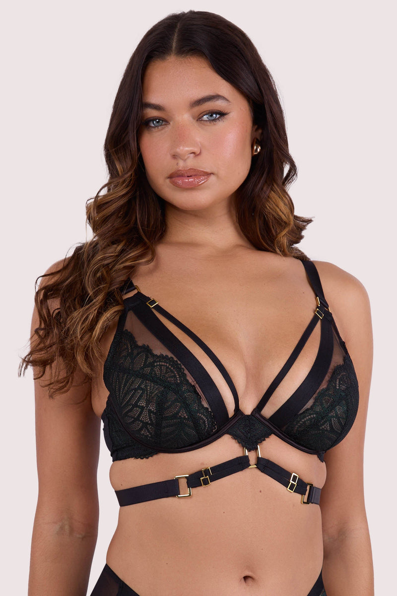 Model wears dark green wet-look plunge bra with underwire cups, strappy harness design, and gold buckle detailing